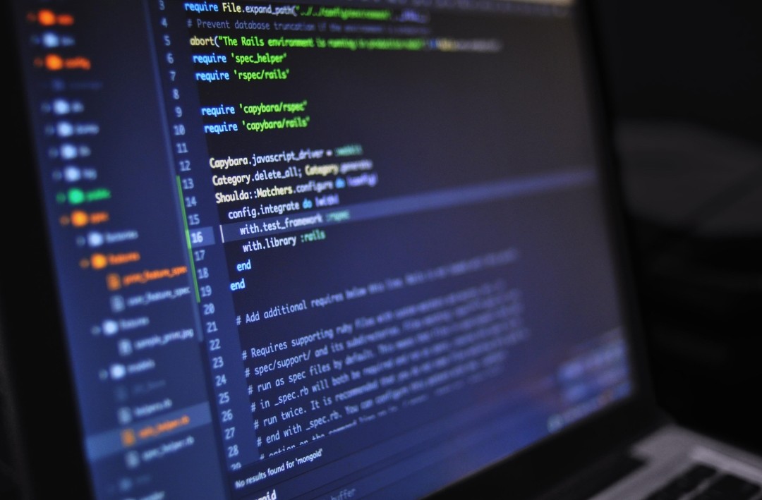 A close-up photograph of a laptop screen displaying lines of code in a dark-themed code editor. The code consists of programming syntax, including highlighted text in green, yellow, and blue. The left sidebar shows a project directory with multiple files and folders. The background is blurred, emphasizing the screen.