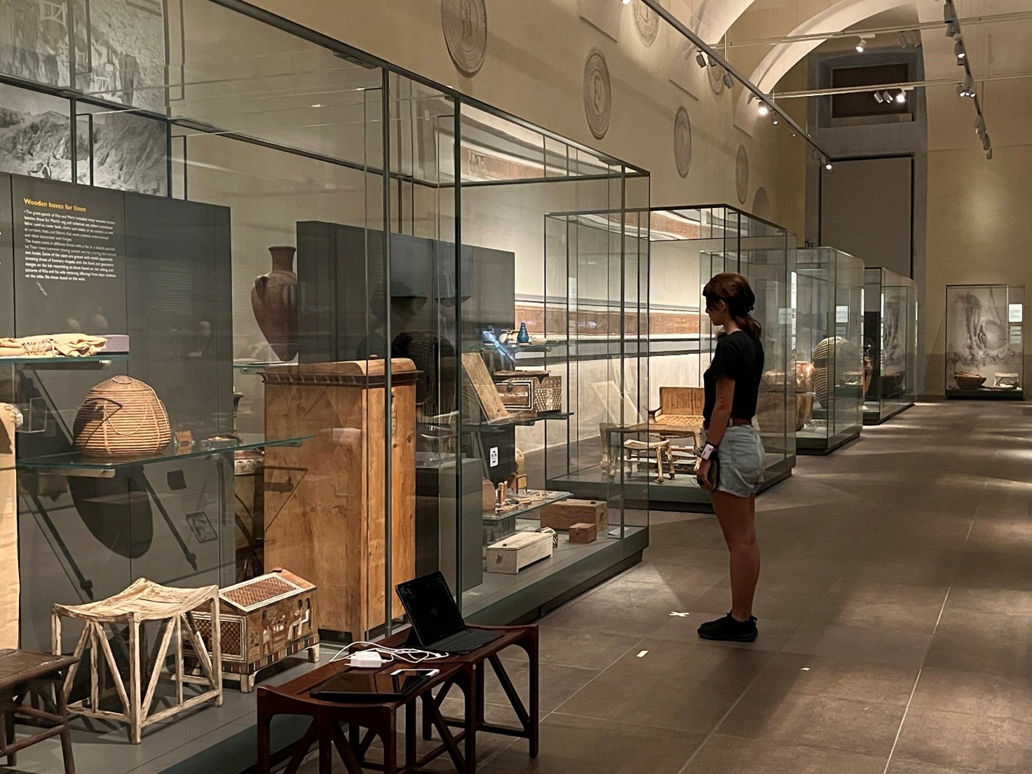 A person stands in a museum gallery, looking at glass display cases filled with artifacts. The gallery has a high arched ceiling and the cases are arranged in a line down the center of the room. A small table with a laptop and other electronic devices is in the foreground.