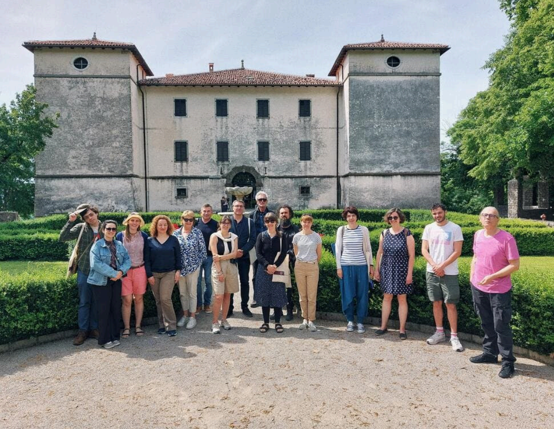 A group of approximately 20 people is standing in a semi-circle on a gravel path in front of a large, old stone building with two towers. The building has a central arched doorway and several small, dark windows. The group, consisting of men and women, is casually dressed, with some wearing hats and sunglasses. Behind them are manicured hedges and trees, and the sky is overcast.