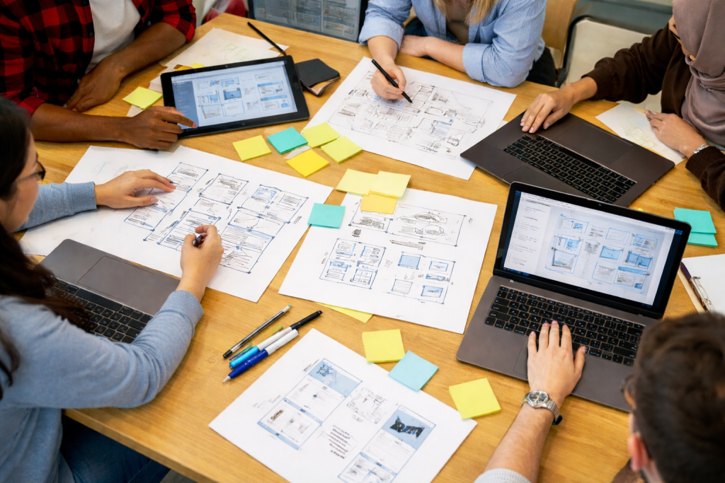 A diverse group of international students working together with researchers in a university lab, reviewing design sketches and digital prototypes on laptops and tablets.
