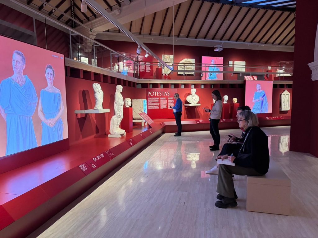 four people sitting in a museum hall on a bank and watching a video screening.