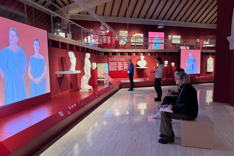 four people sitting in a museum hall on a bank and watching a video screening.