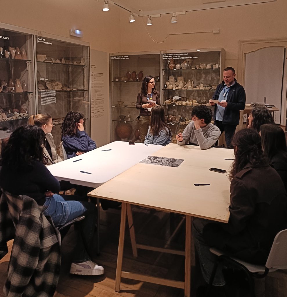 a group of people involved in a co-creation activity within a room. five of them are sitting on chairs around the table, and two of them are presenting to the group. All others who are sitting are watching them.,